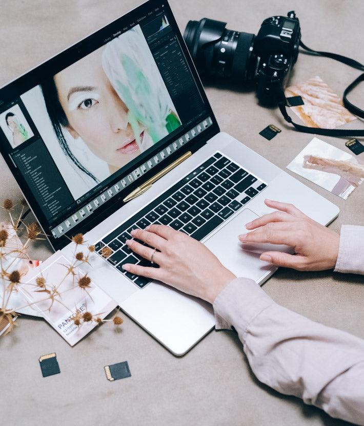 person using laptop to retouch photo of a woman standing next to a flower. SD cards, photos and her camera surround the laptop while the woman works on retouching the photos on her laptop