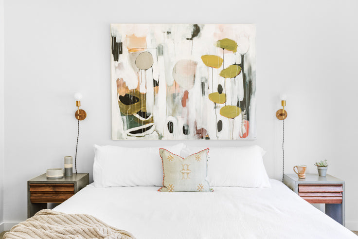 custom pink and green abstract painting hung up above white bed with a tan throw blanket two colourful nightstands surround the bed and hole a mug and flower pot on them