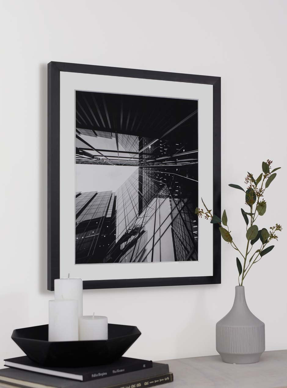 modern black frame called the alma that is 1/2 inch wide facing the front with a white mat and a black and white photograph of a reflective new york city building above a table with a grey vase holding green leaves and a modern dish on top of coffee table books with white candles.