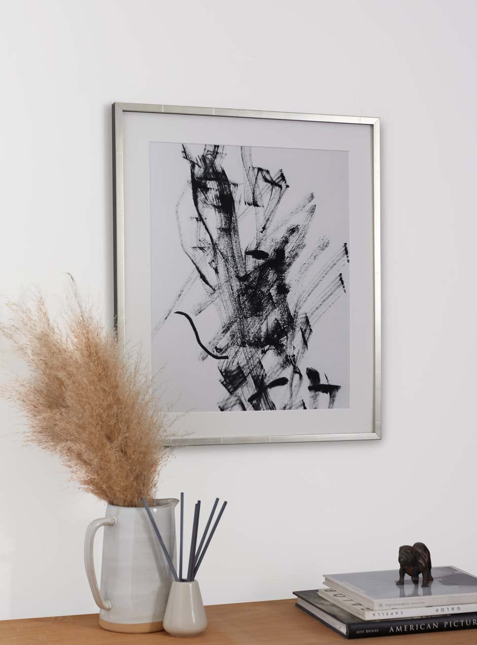 reflective metallic silver thin frame with various lines across moulding shown with white mat and an abstract ink line art piece above a table with a fluffy dried plant and an incense diffuser in a high end apartment