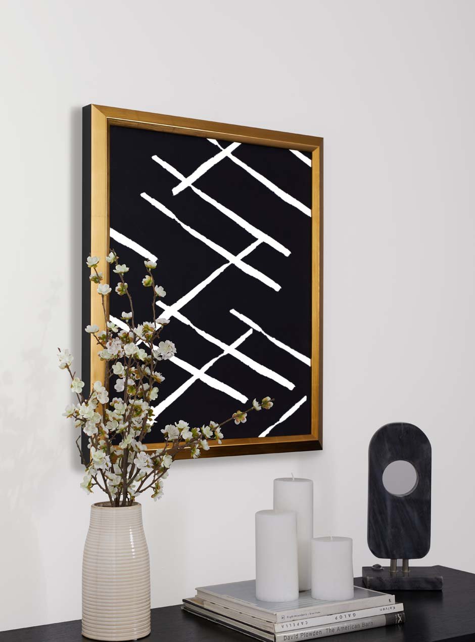 bright yellow gold leafed frame with bevel towards the center artwork which is dark black with white criss cross pattern modern art on white wall with white flowers in vase in front of gold frame and white candles with modern decor and books