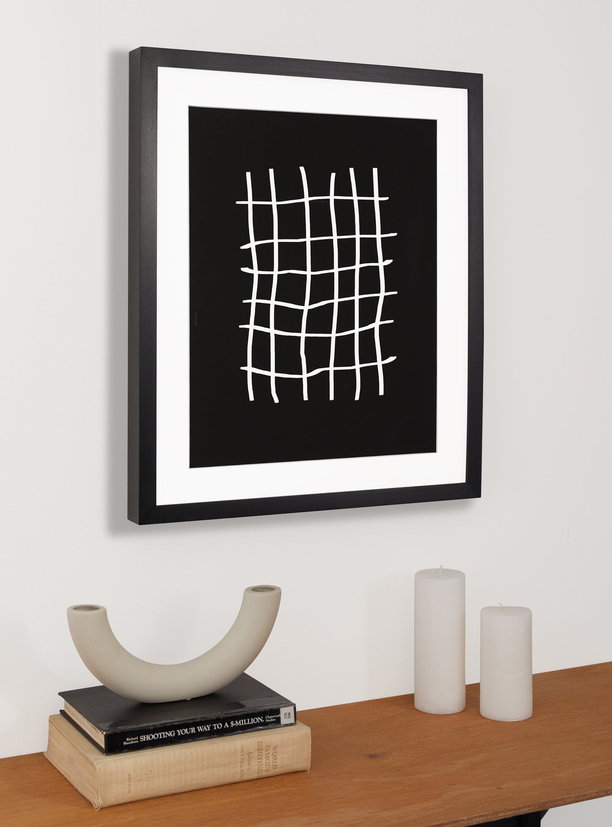 Photograph of a frame in a home with a table underneath showing various itemsblack solid maple frame with white matboard and black and white abstract art resembling a pound sign multiple times