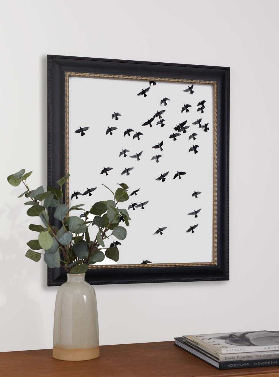 birds flying with white background in black frame and gold inner trim above table with potted branches and magazine stack
