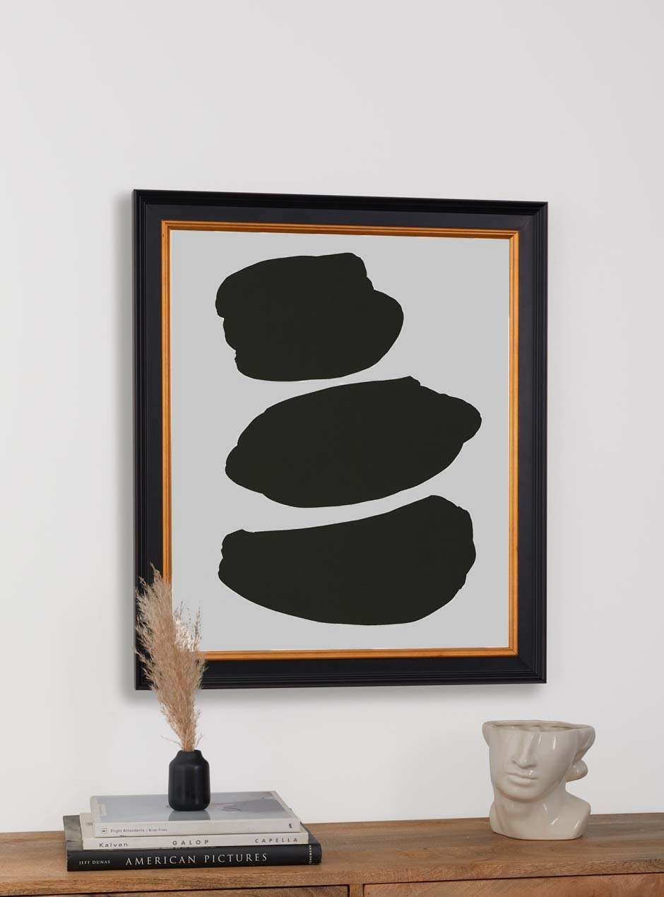 3 abstract black shapes on a white background in a black and gold frame above a table with books and sculpture of half a face