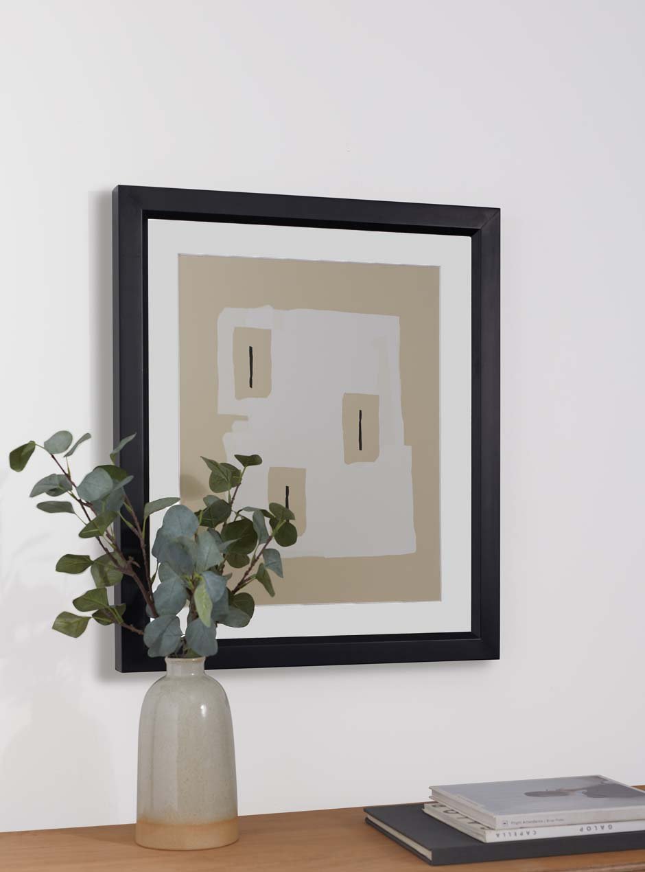 black frame with slight bevel towards center showcasing brown and neutral artwork with white mat on a white wall with eucalyptus plant in a beige vase on the table slightly obstructing the high end quality frame and books