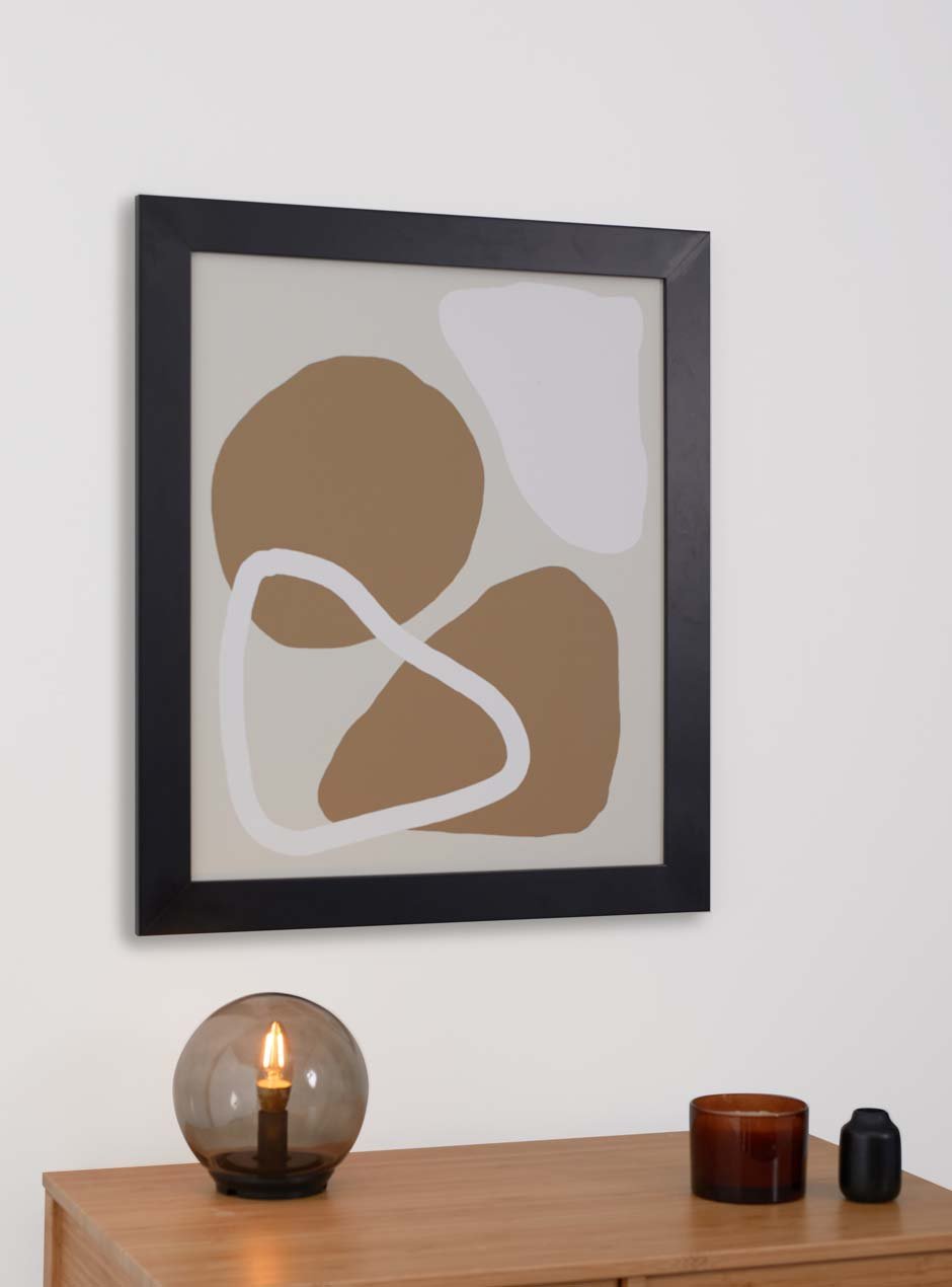 black skyframe wide frame called agbes with neutral tone abstract artwork inside hanging on plain wall modern style with wooden desk in front with spherical light and candles